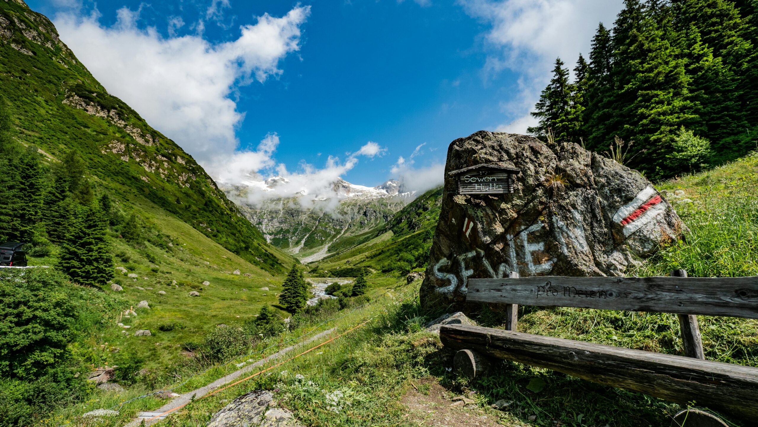 A picturesque view of a Swiss valley with a wooden bench, trail marker, and mountain backdrop, perfect for hikers.